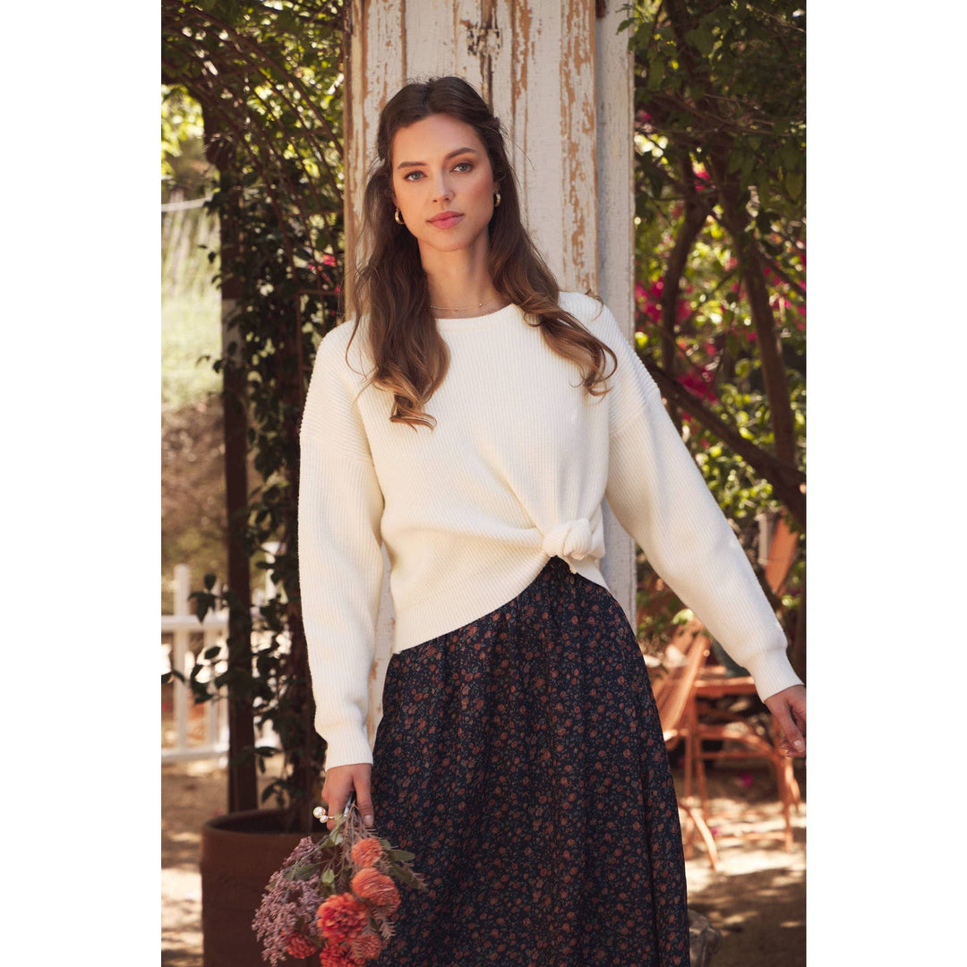 Model wearing Bow Back Sweater | Dolman Sleeve Knit with Satin Tie. Lifestyle photo with outdoor garden setting.  Model leaning with arm out showing another angel.