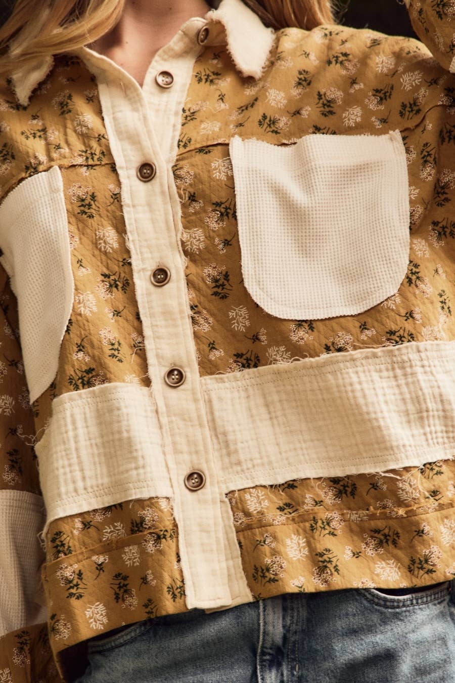 A close-up of a person wearing a patchwork-style button-front jacket in warm tan and cream fabrics with a small floral print. The jacket features solid cream pocket panels and trim, brown buttons, and blue jeans visible at the bottom.