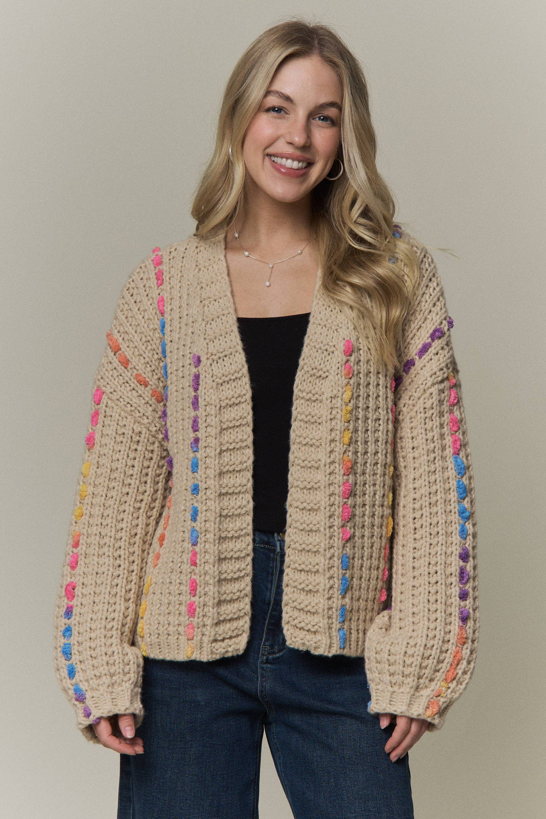 A woman wearing a chunky knit cardigan in beige with colorful vertical yarn stripes in pink, blue, yellow, orange, and purple, styled with a black top and blue jeans against a solid light gray background.