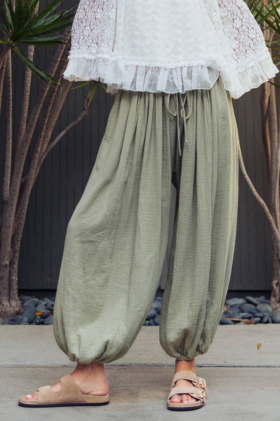 Woman wearing olive green crinkle cotton balloon pants with drawstring waist and elastic ankle cuffs paired with a white lace blouse.
