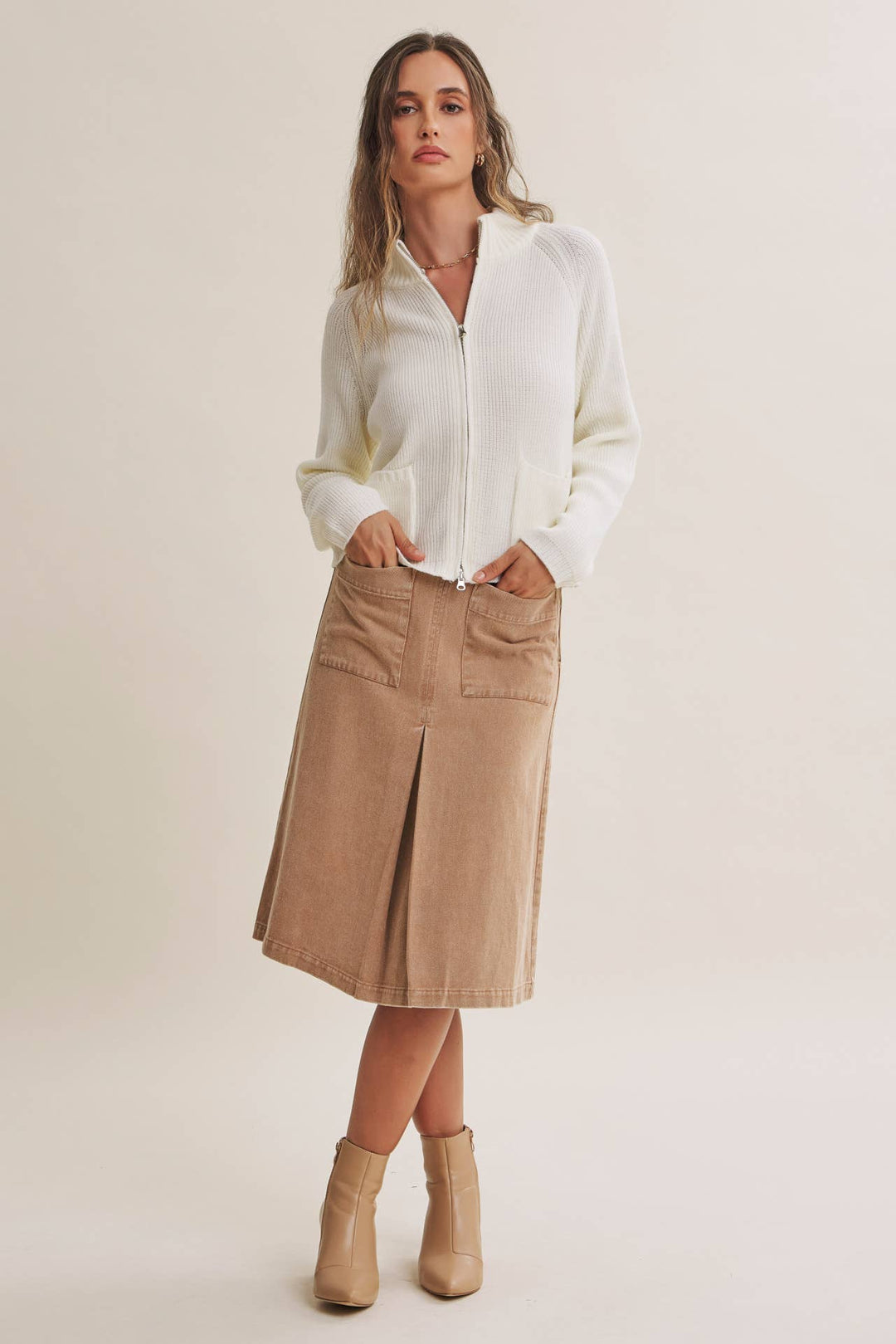 A woman modeling a white long-sleeve zip-front blouse and a beige knee-length pleated skirt with front pockets, paired with beige ankle boots, against a plain light beige studio background.