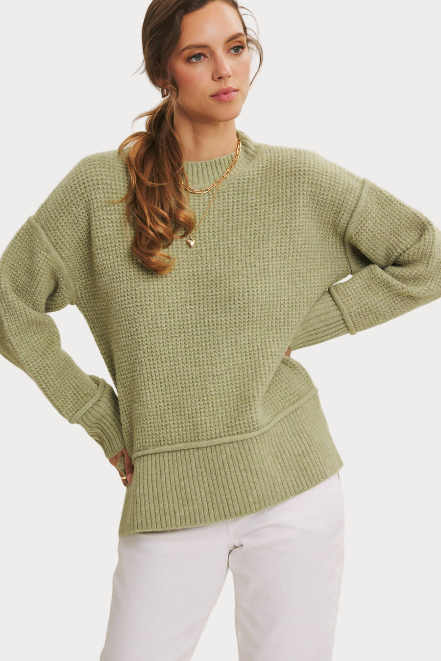 Woman wearing a light sage green recycled blend knit sweater with ribbed trim, styled casually with white pants.
