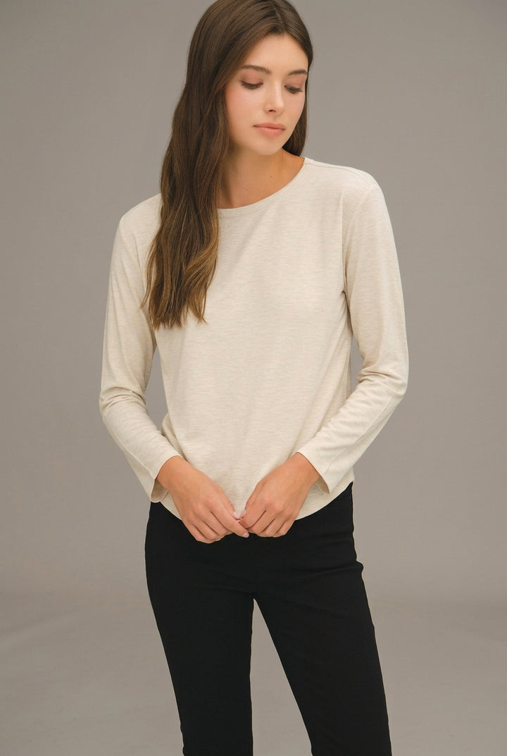 A woman wearing a cream-colored long-sleeve crewneck top and black pants, standing against a neutral gray studio background.