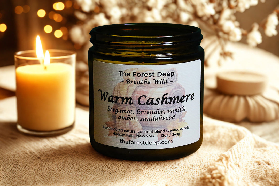 Candle jar labeled 'Warm Cashmere' by The Forest Deep with a lit candle in the background.