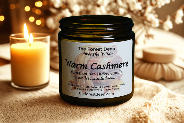 Candle jar labeled 'Warm Cashmere' by The Forest Deep with a lit candle in the background.