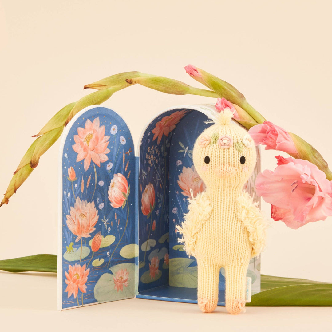 Knitted duckling toy standing next to a book with floral illustrations, surrounded by pink flowers and green leaves.