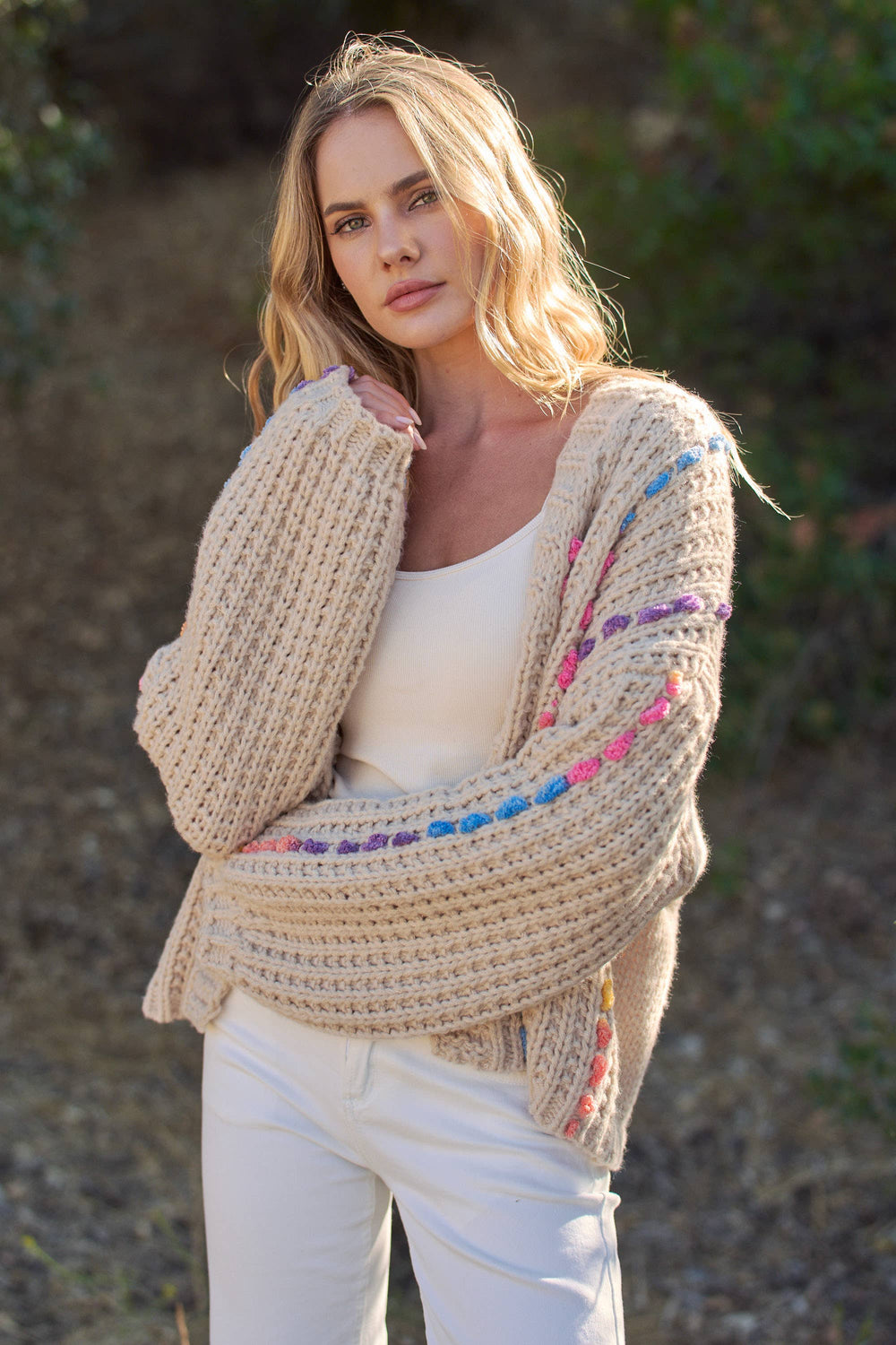 A woman wearing a chunky knit cardigan with colorful crochet accents, styled over a white tank top and white pants in an outdoor setting.