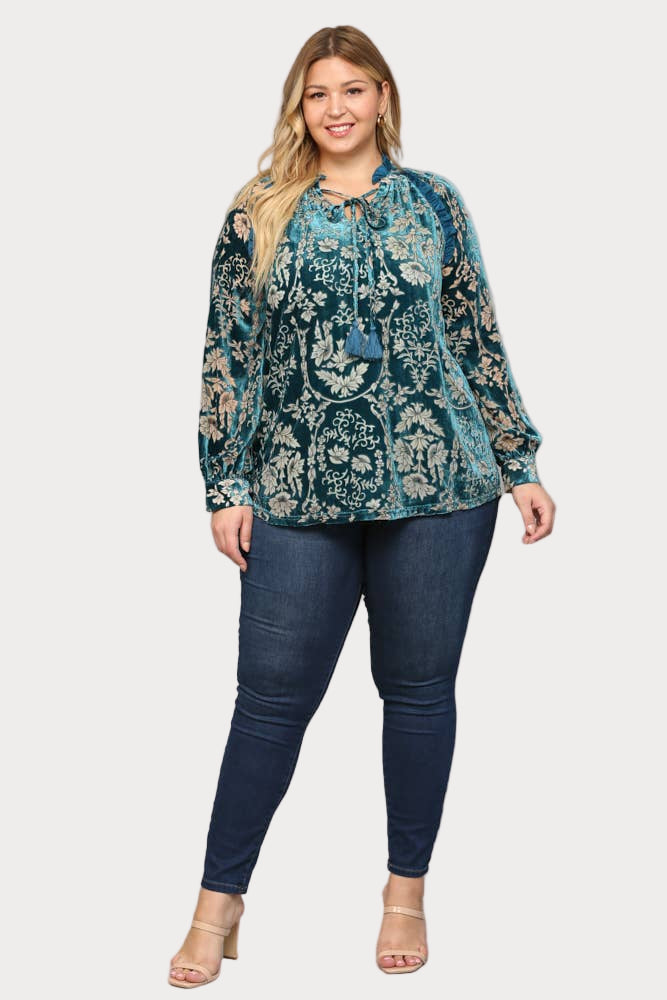 A smiling plus-size model wears a long-sleeve teal burnout velvet blouse featuring a floral pattern, ruffle trim, and a tassel tie at the neck.

