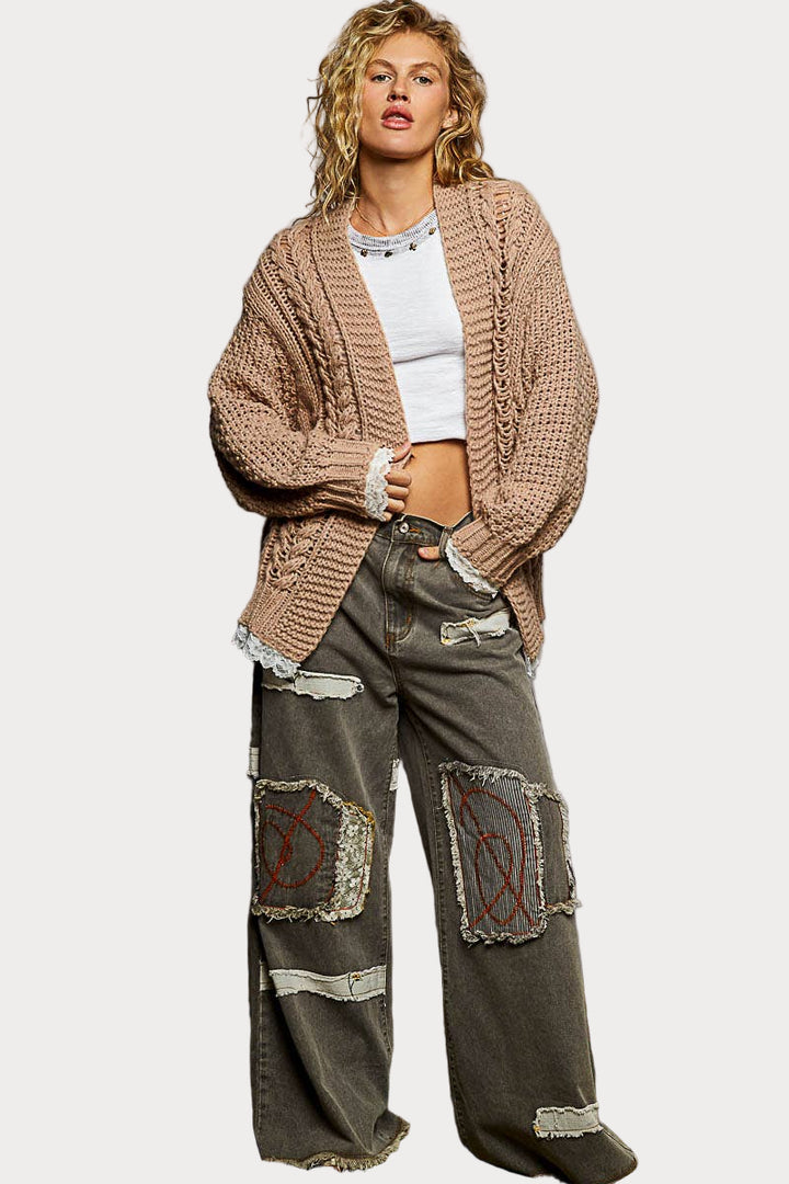 A woman models a beige, chunky cable knit cardigan featuring an open front and delicate white lace detailing on the hem and sleeve cuffs.
