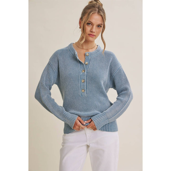 A woman wearing a light blue ribbed knit sweater with a buttoned henley neckline and white pants, standing against a plain beige studio background.