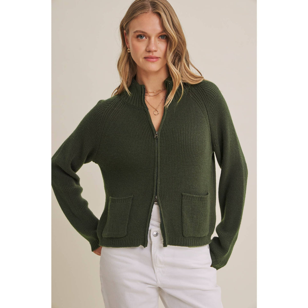 Woman wearing a green zip-up sweater with white pants against a neutral background