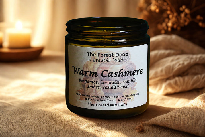 Candle jar labeled 'Warm Cashmere' by 'The Forest Deep' on a soft surface.