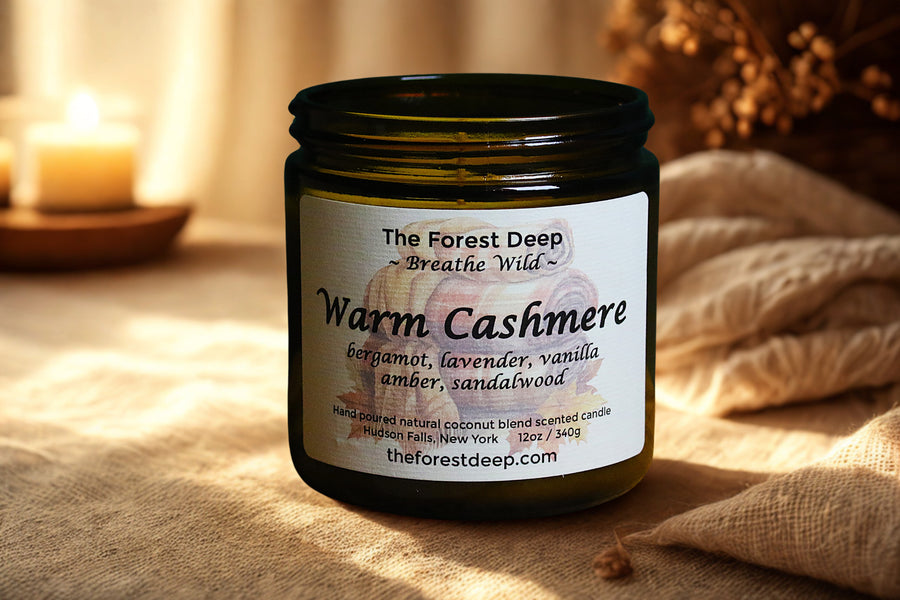 Candle jar labeled 'Warm Cashmere' by 'The Forest Deep' on a soft surface.
