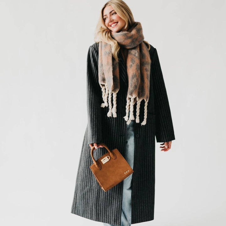 Woman wearing a long coat and patterned scarf with a brown handbag on a plain background