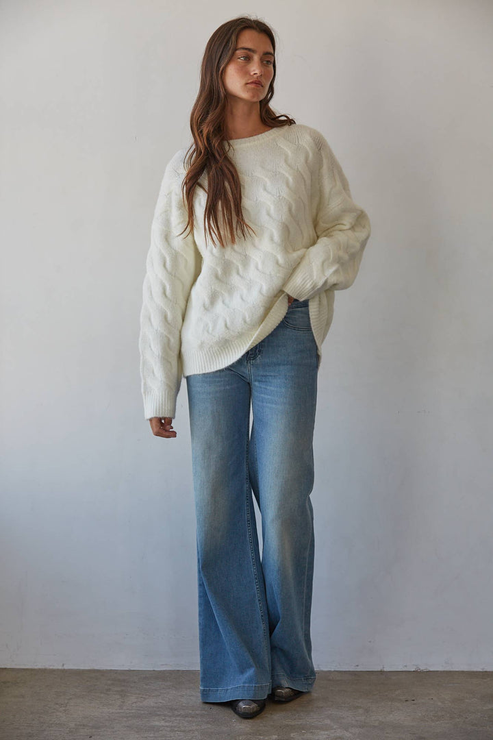 A woman modeling a cream cable-knit sweater and wide-leg blue jeans against a plain light gray wall.