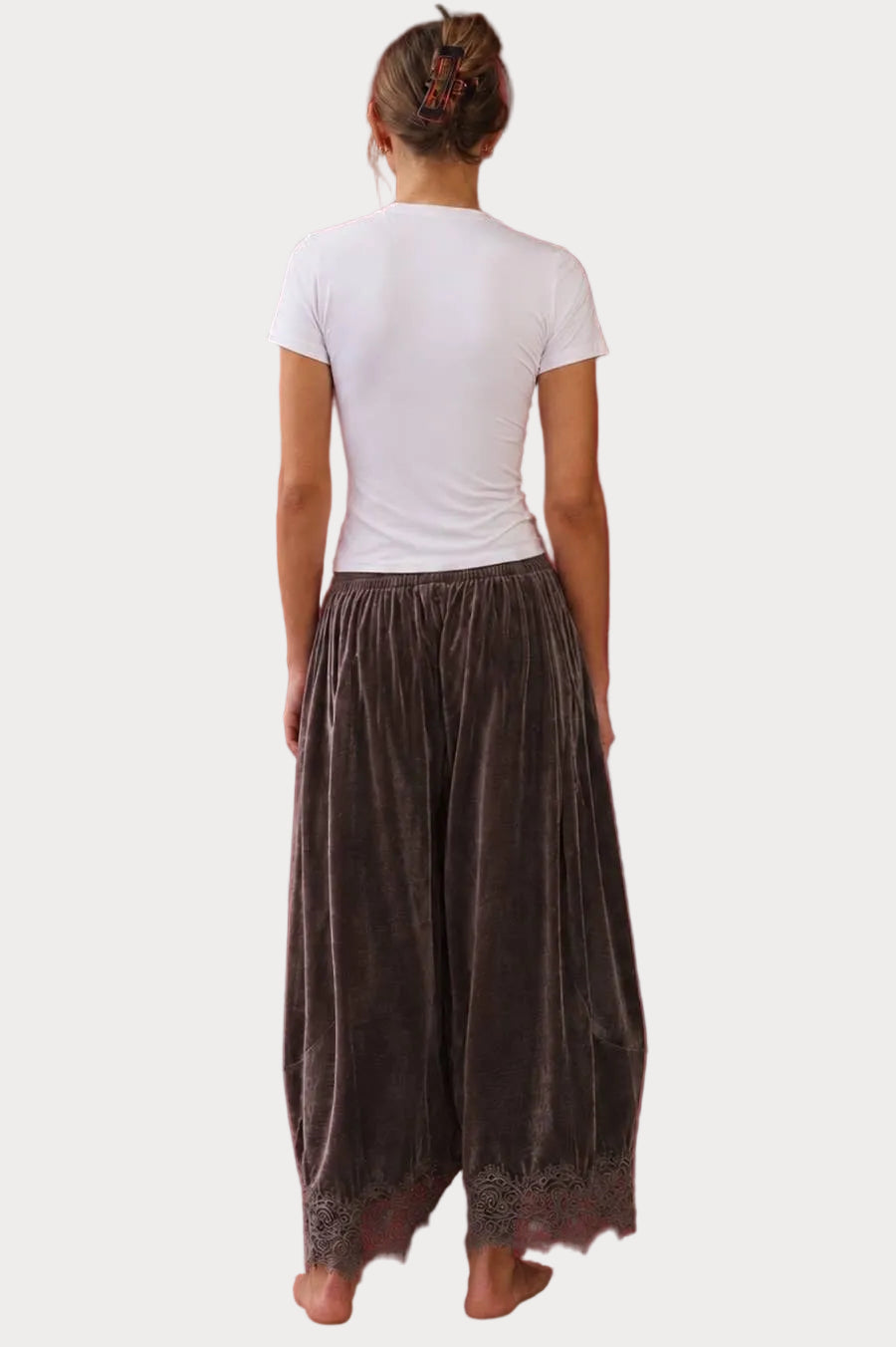 A woman viewed from behind wearing a white short-sleeve t-shirt and dark brown velvet harem pants with a lace hem, standing against a solid background.