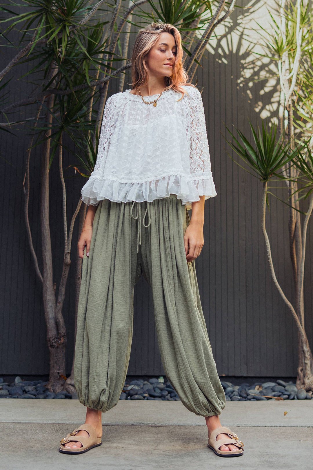 Woman wearing olive green crinkle cotton balloon pants with drawstring waist and elastic ankle cuffs paired with a white lace blouse.