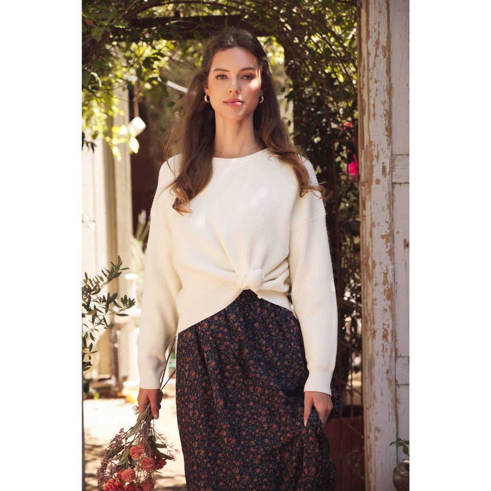 Model wearing Bow Back Sweater | Dolman Sleeve Knit with Satin Tie. Lifestyle photo with outdoor garden setting. Model facing forward holding skirt. Sweater styed with a knot at the waist.