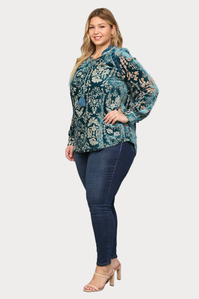 A smiling plus-size model wears a long-sleeve teal burnout velvet blouse featuring a floral pattern, ruffle trim, and a tassel tie at the neck.

