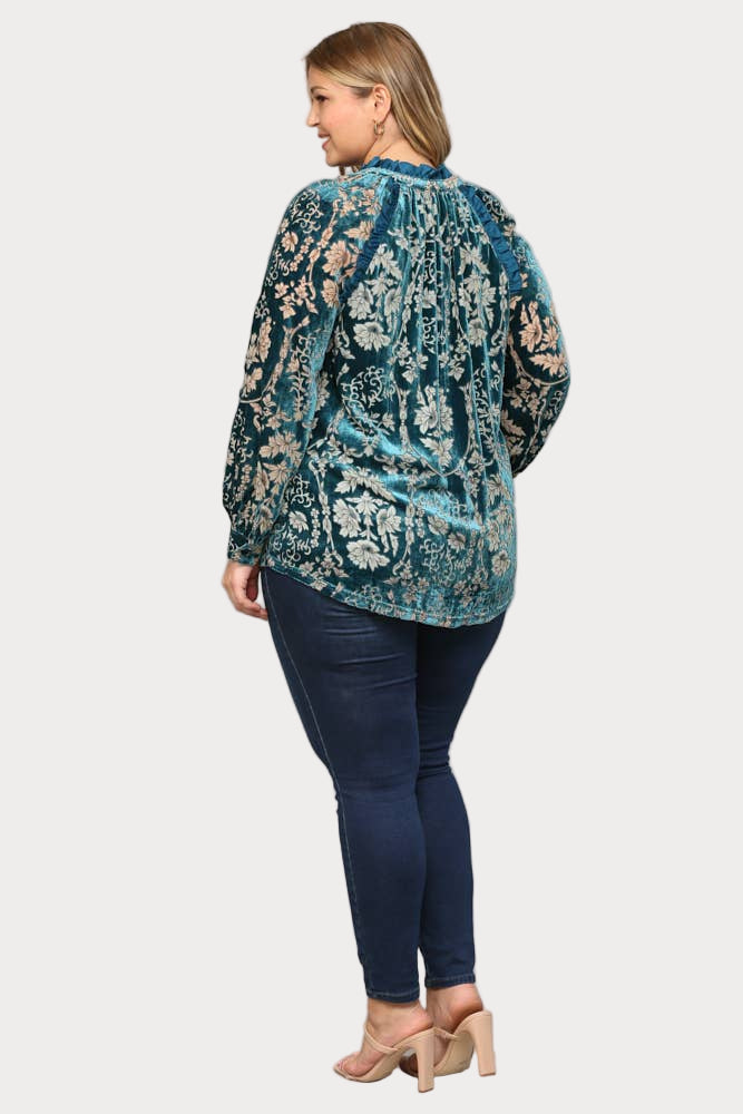 A smiling plus-size model wears a long-sleeve teal burnout velvet blouse featuring a floral pattern, ruffle trim, and a tassel tie at the neck.

