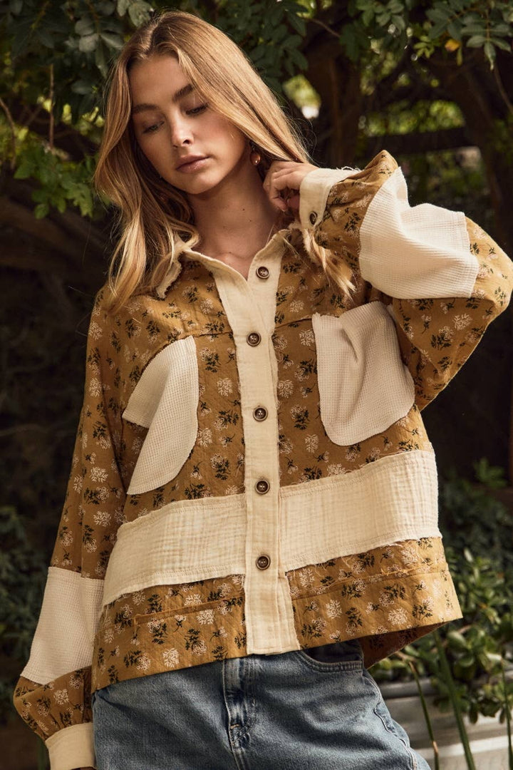 A woman wearing a patchwork-style button-front jacket with a tan floral print and cream panels, paired with blue jeans, photographed outdoors in front of leafy trees.