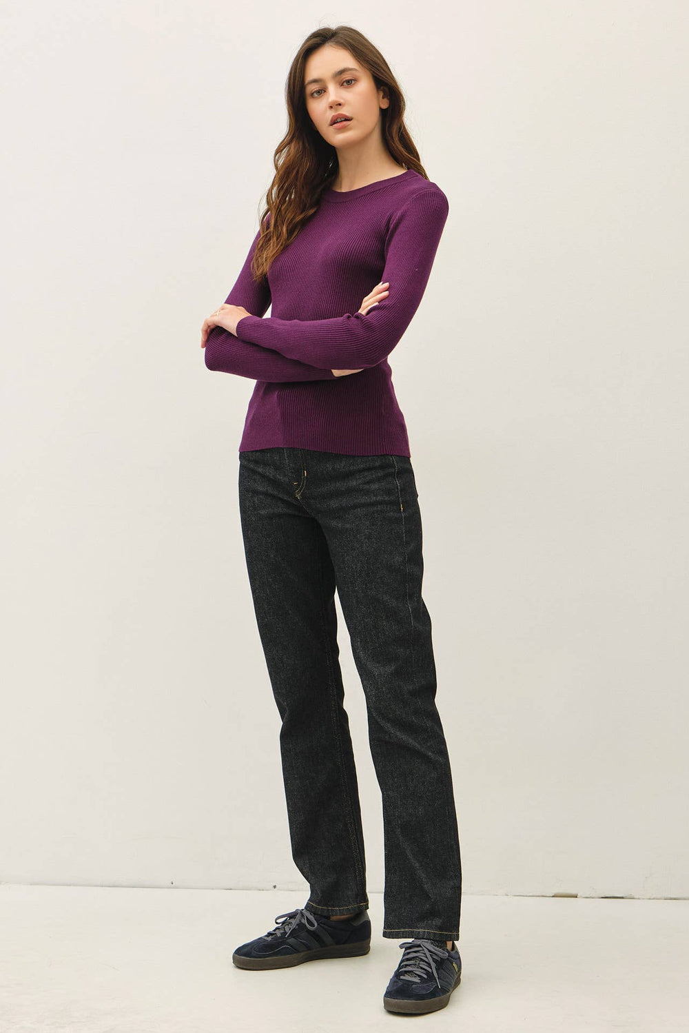 A woman modeling a long-sleeve crewneck knit sweater in deep purple, paired with dark gray jeans and dark blue sneakers, standing against a plain off-white studio background.