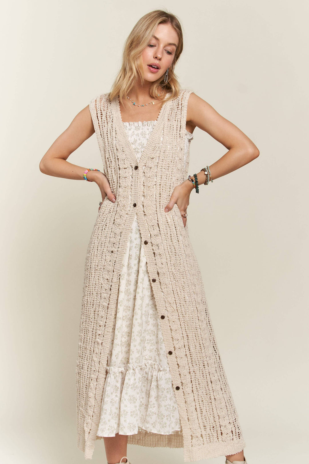 Woman wearing long beige crochet duster vest over a white floral dress
