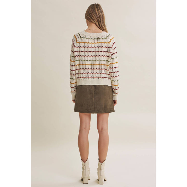 A woman shown from the back wearing a cream knit sweater with horizontal multicolor stripes and a brown mini skirt, paired with white ankle boots, against a plain beige studio background.
