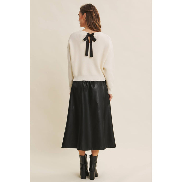Woman wearing a Bow Back Sweater | Dolman Sleeve Knit with Satin Tie paired with a faux black leather skirt on a beige background. Model showing back detail of the satin bow on the back of sweater.