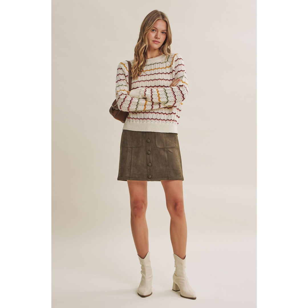 A model stands against a plain beige studio background wearing a cream knit sweater with multicolor horizontal stripes, a dark brown mini skirt with front buttons, and off-white ankle boots.