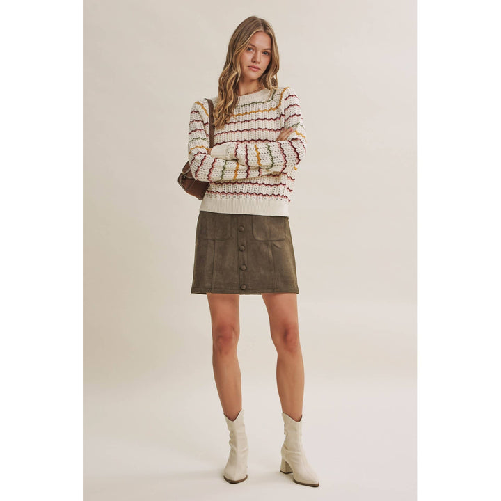 A model stands against a plain beige studio background wearing a cream knit sweater with multicolor horizontal stripes, a dark brown mini skirt with front buttons, and off-white ankle boots.
