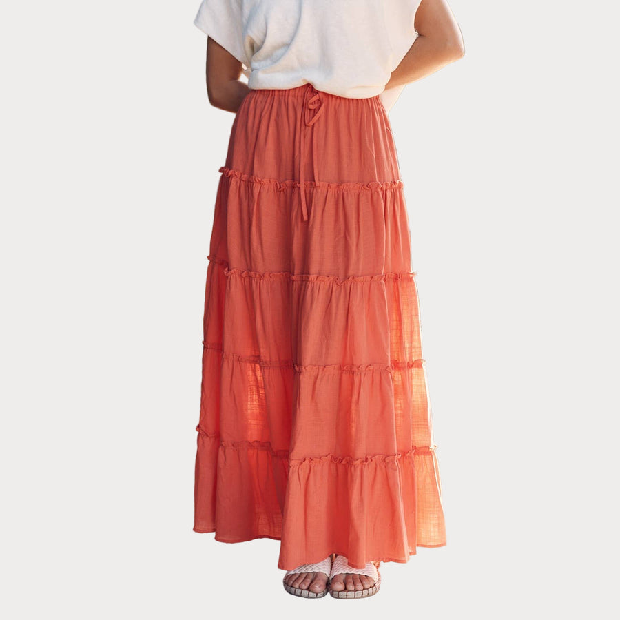 A person wearing a tiered ruffled maxi skirt with an elastic waistband, in an orange color, paired with a white top, standing outdoors.