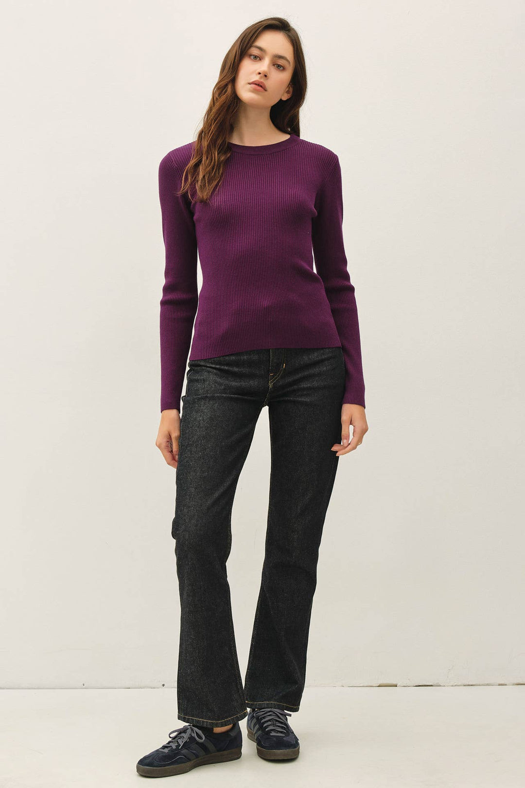 A woman modeling a long-sleeve crewneck sweater in deep purple, paired with dark wash jeans and navy sneakers, standing against a plain off-white studio background.