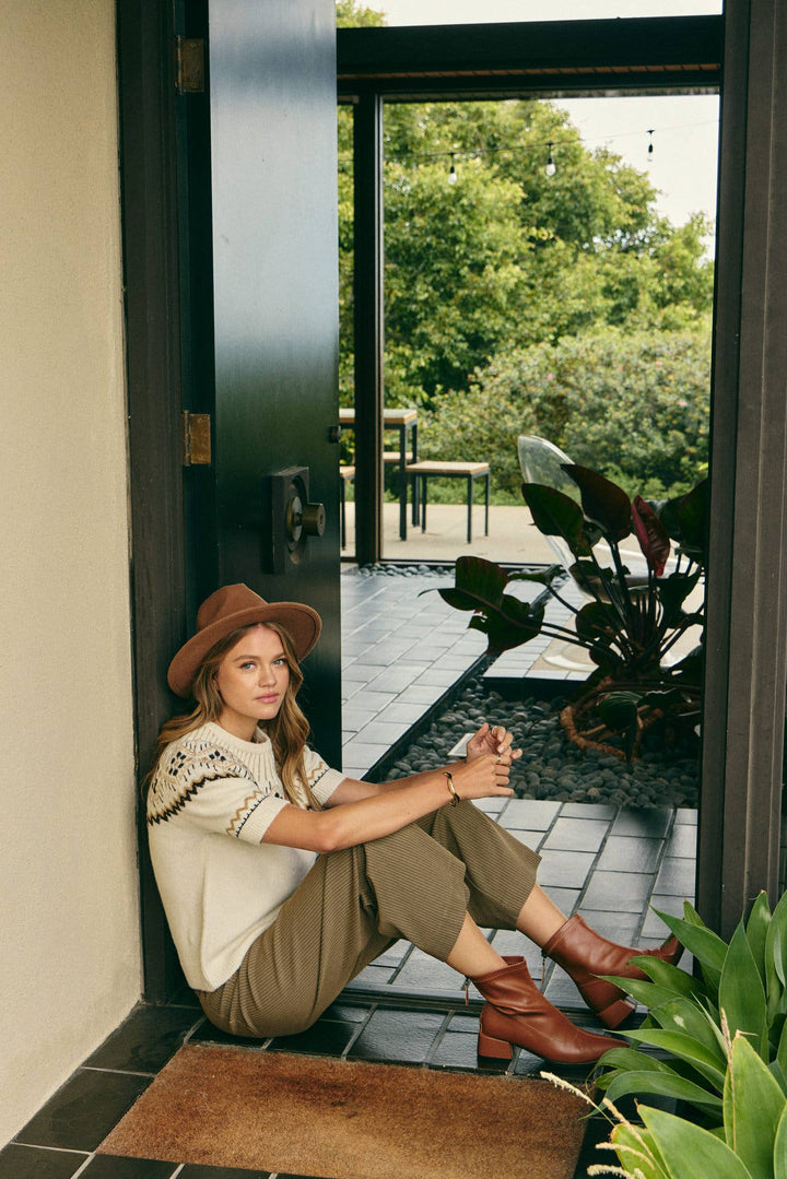 A woman sits on a tiled doorstep wearing a brown wide-brim hat, a cream knit top, olive-brown cropped pants, and brown ankle boots, with a garden visible outside.