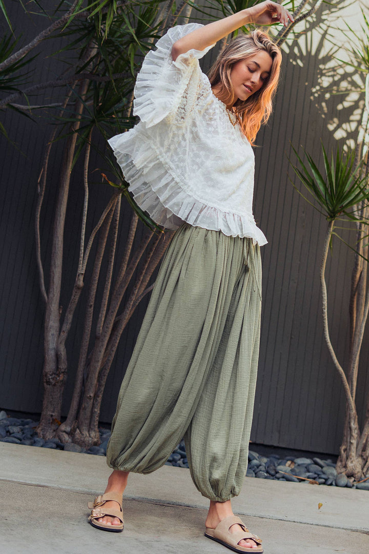 Woman wearing olive green crinkle cotton balloon pants with drawstring waist and elastic ankle cuffs paired with a white lace blouse.