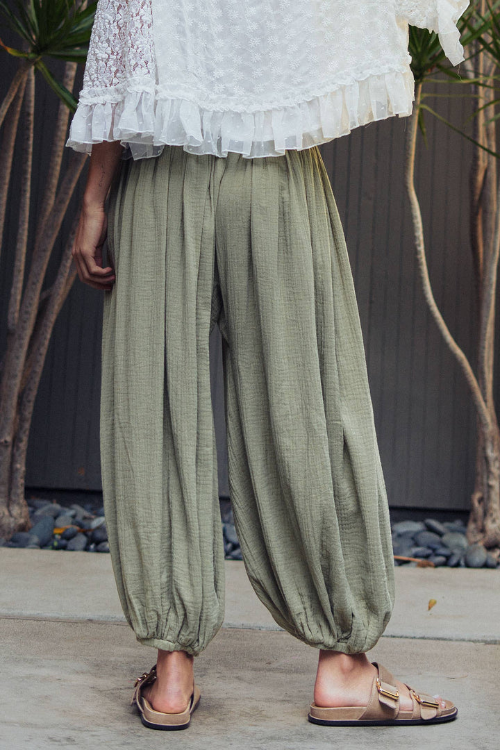 Woman wearing olive green crinkle cotton balloon pants with drawstring waist and elastic ankle cuffs paired with a white lace blouse.