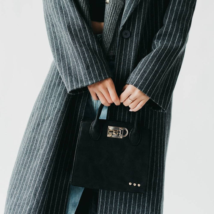 Person wearing a long pinstripe coat holding a black handbag with a gold clasp on a light gray background
