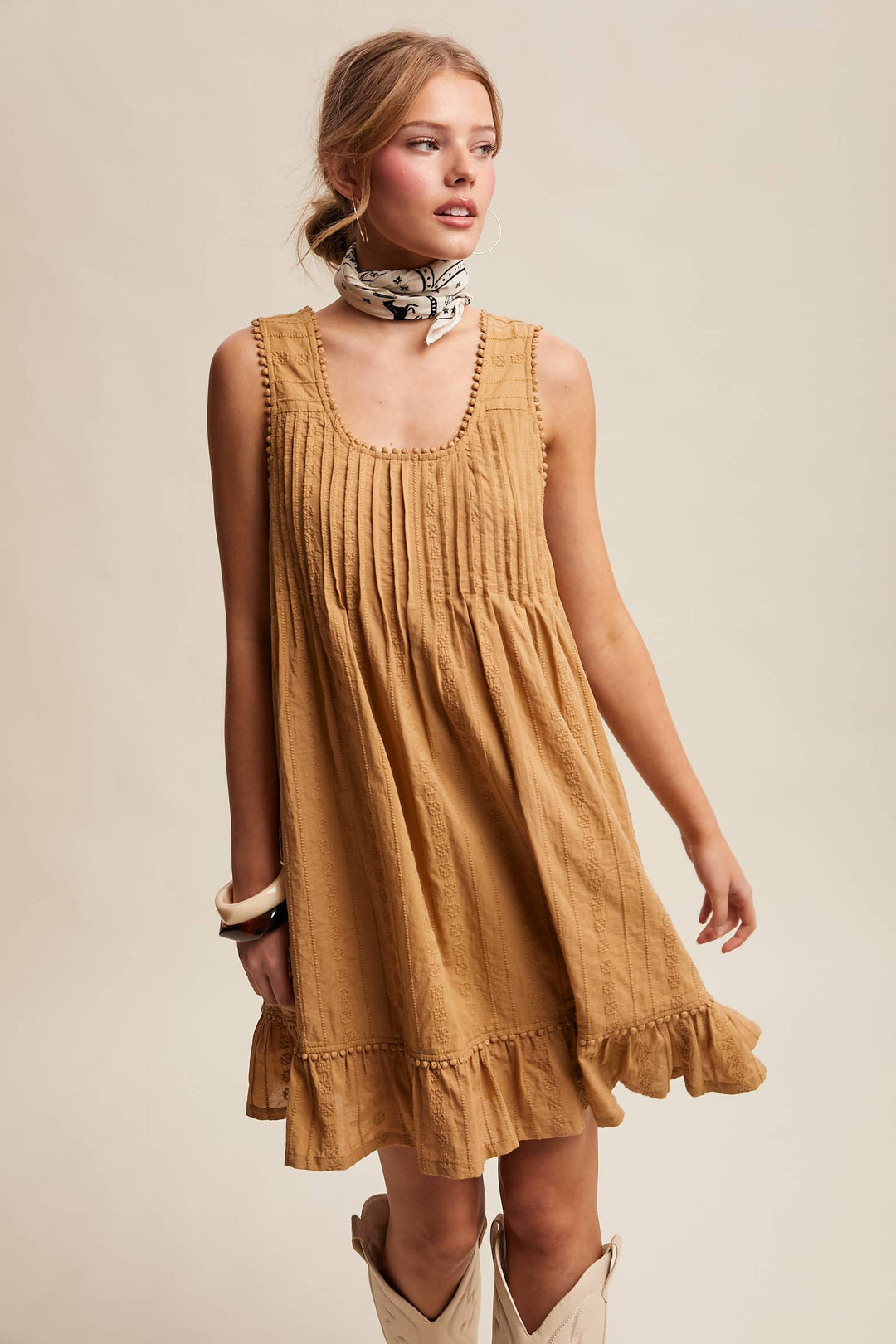Woman wearing a textured camel-colored sleeveless mini dress with pintuck details and ruffle hem.