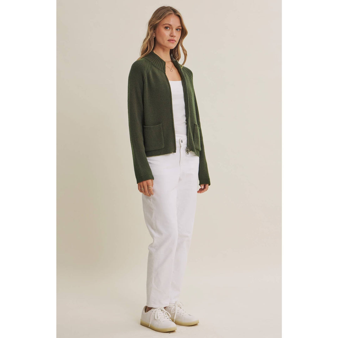 Woman wearing a sweater green jacket, white top, and white pants on a beige background