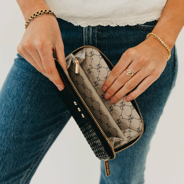 Person holding a black clutch wallet  with gold accents, wearing blue jeans and a white top.