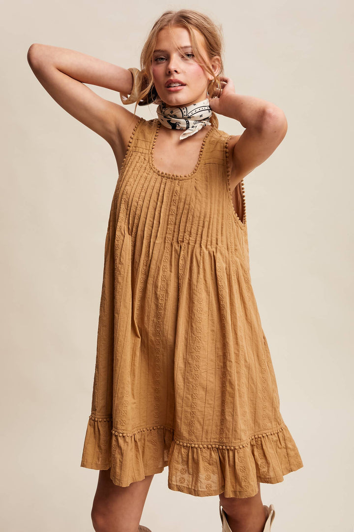 Woman wearing a textured camel-colored sleeveless mini dress with pintuck details and ruffle hem.
