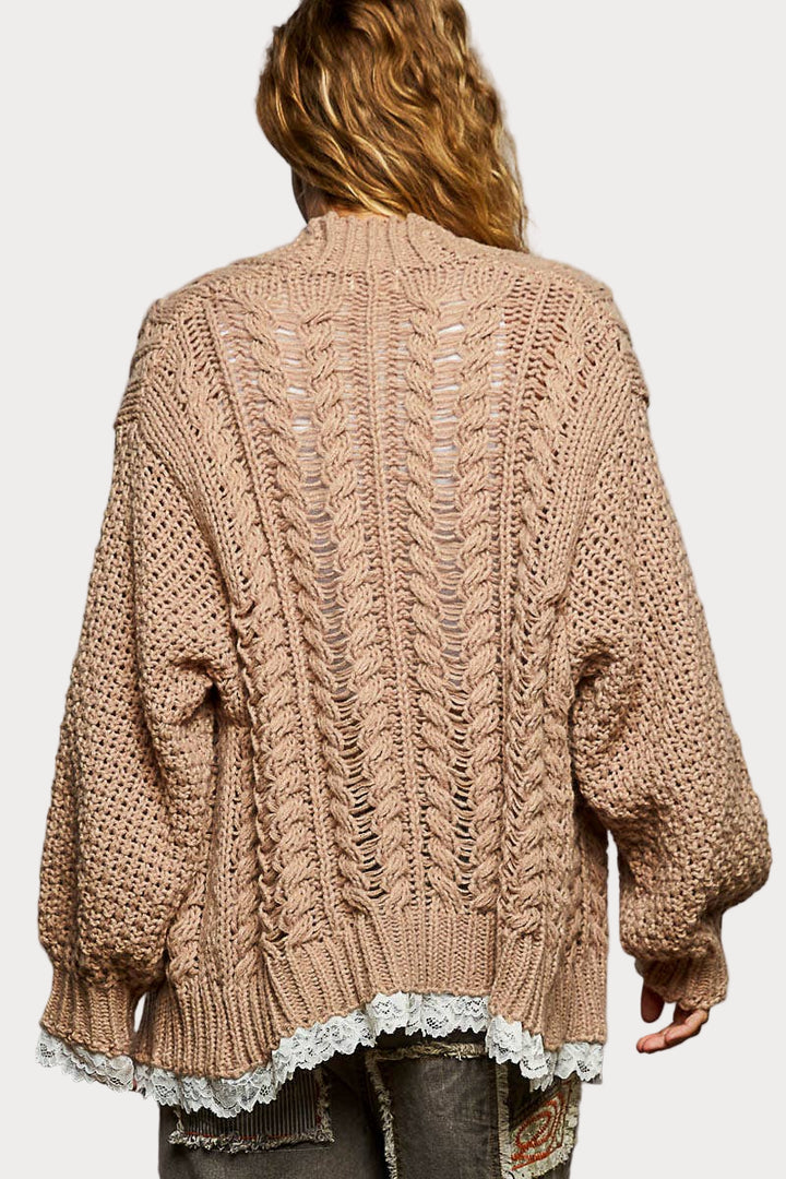 A woman models a beige, chunky cable knit cardigan featuring an open front and delicate white lace detailing on the hem and sleeve cuffs.
