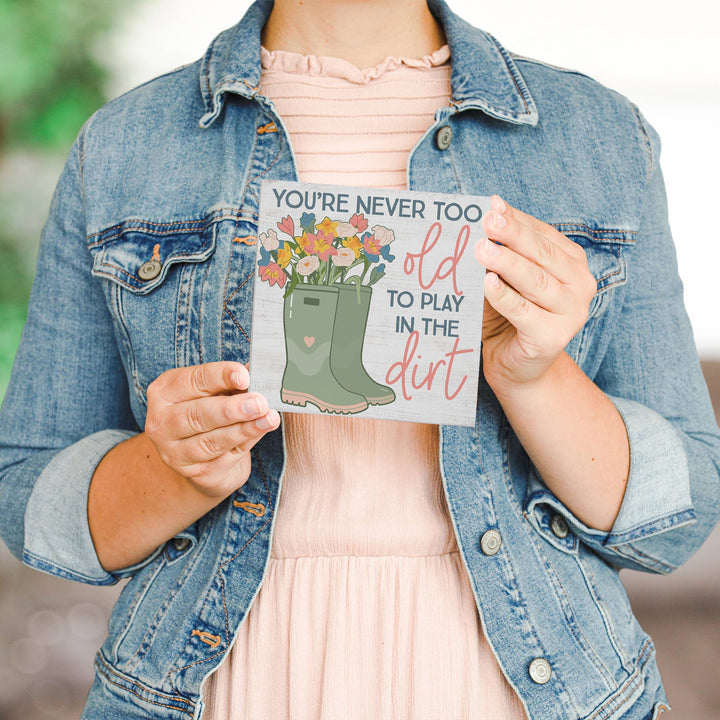 Person holding a decotative sign with floral design and text, "You're never too old to play om the dirt", wearing a denim jacket and pink dress.
