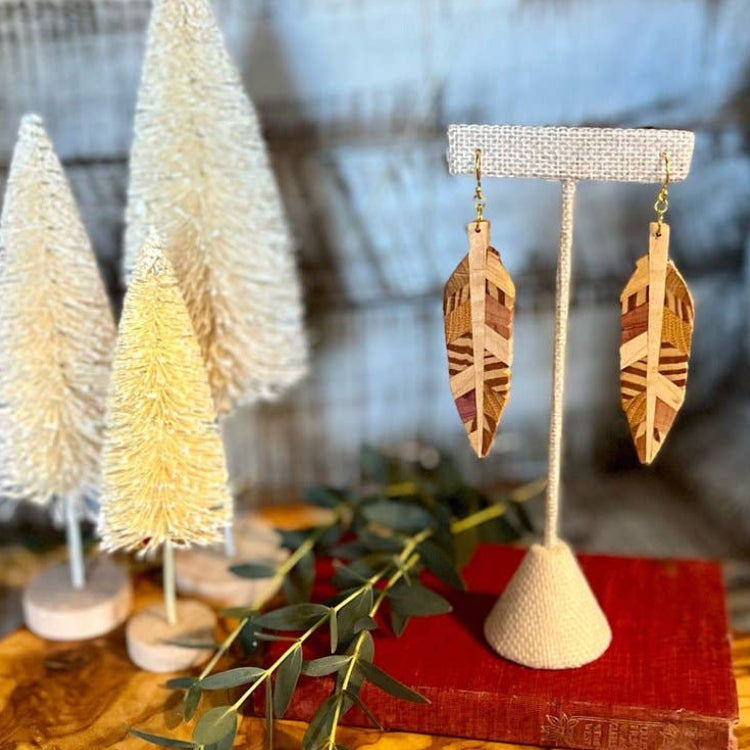 Wooden feather earrings on a stand with decorative trees and greenery in the background.