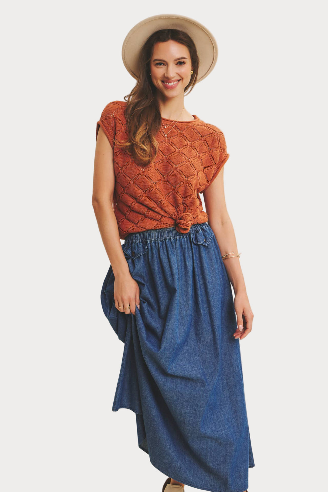 Woman wearing a rust-colored top and blue skirt with a beige hat on a plain background