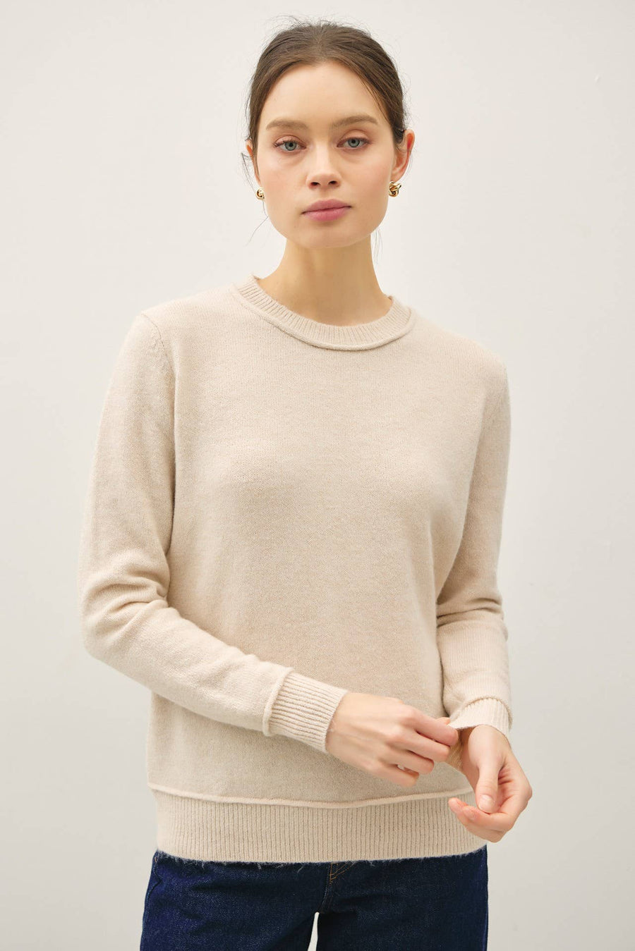 A woman wearing a light beige crewneck knit sweater and dark blue jeans, standing against a plain light gray studio background.