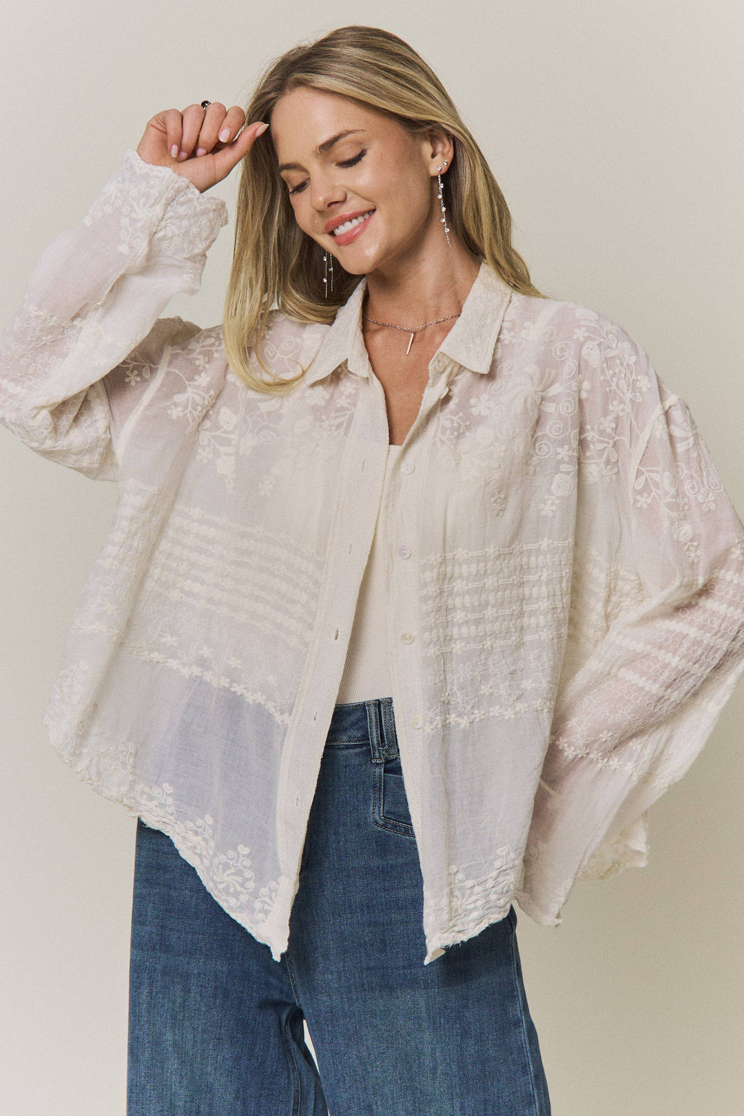 Woman wearing a light-colored lace embroidered blouse and blue jeans against a plain background
