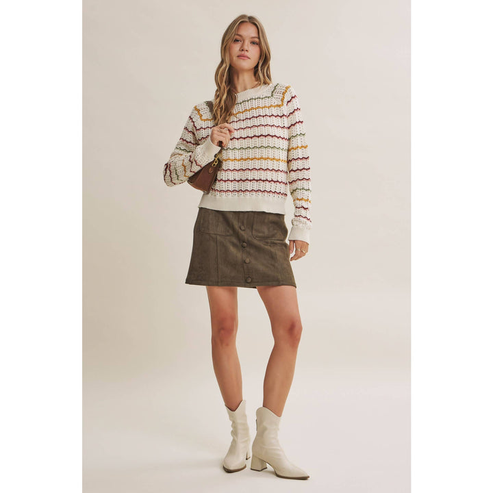 A woman modeling a cream knit sweater with multicolor horizontal stripes, paired with an olive green mini skirt and white heeled boots, against a solid light beige studio background.