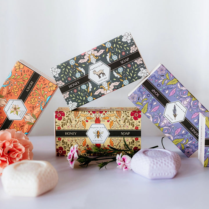Four decorative honey soap gift boxes arranged on a white surface with two textured honey bars in front. The boxes feature colorful floral and nature illustrations with black bands labeled 'HONEY SOAP' and 'BEEWAX SOAP'.