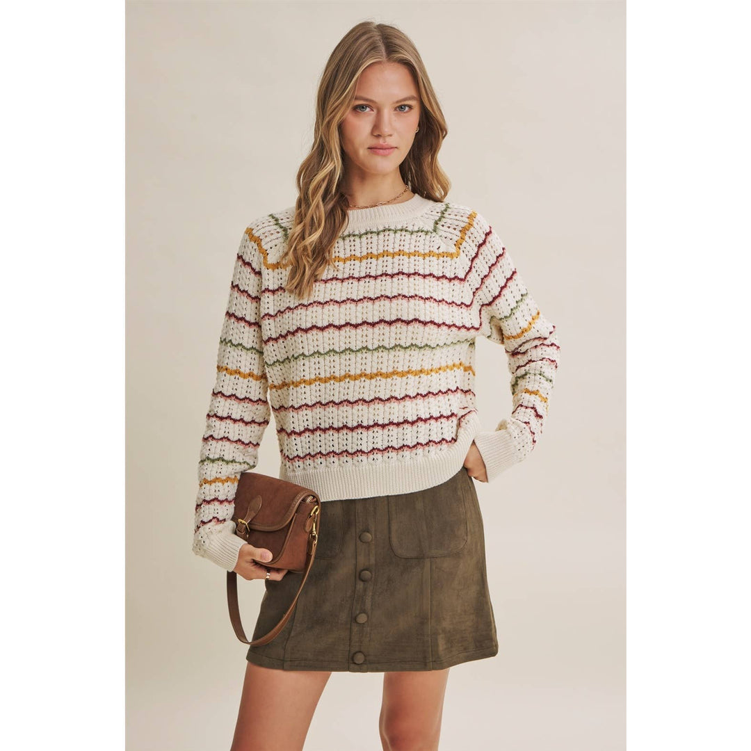 A woman modeling a cream knit sweater with thin multicolor horizontal stripes in red, yellow, and green, paired with an olive green skirt and holding a brown crossbody bag against a plain beige studio background.